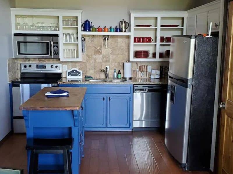 Kitchen an Oklahoma cabin rental with blue cabinets, stainless steel appliances, a small island with seating, and open shelves displaying dishes and glassware.