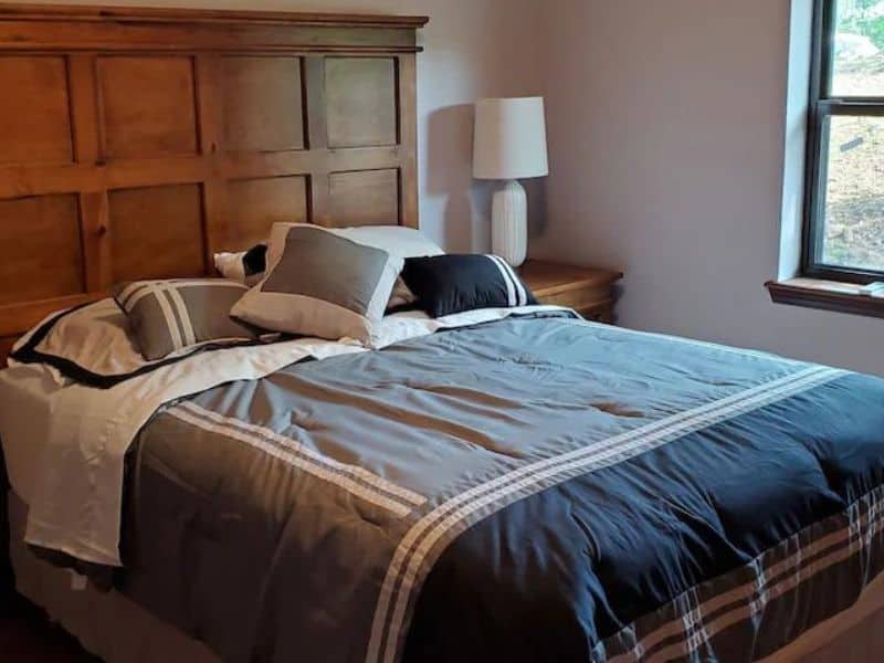 Bedroom with a wooden headboard, a bed with gray bedding, pillows, a nightstand, and a window.