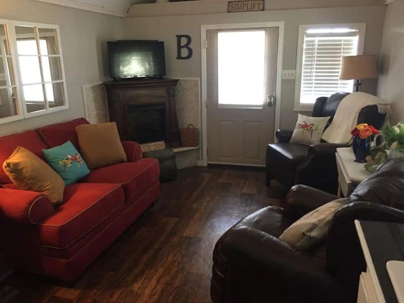 Cozy living room in a tiny house Vrbo near Turner Falls, Oklahoma featuring a red sofa, leather chairs, a fireplace, and a TV.