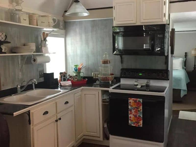 Small kitchen in a tiny house vacation rental near Turner Falls, Oklahoma with white cabinets, a stove, microwave, and open shelving.