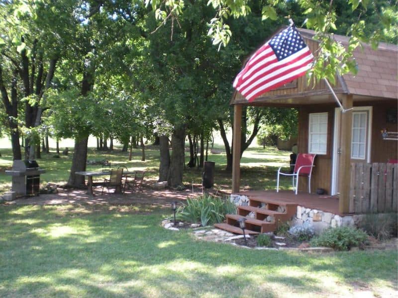 Exterior view of a tiny house rental near Turner Falls, Oklahoma with an American flag, outdoor seating, and a barbecue area surrounded by trees.