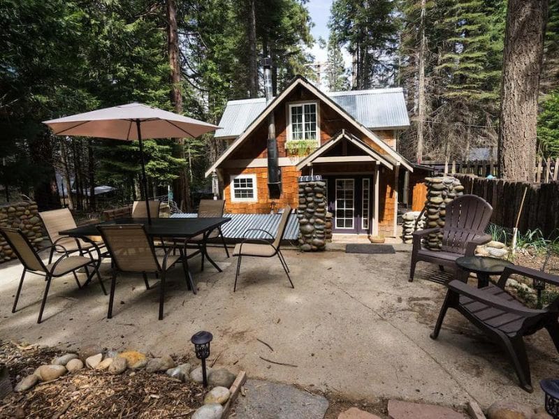 A cozy, rustic cabin among tall pine trees, featuring a spacious outdoor patio with a dining table, chairs, and a large umbrella. A pet-friendly cabin rental near Yosemite.