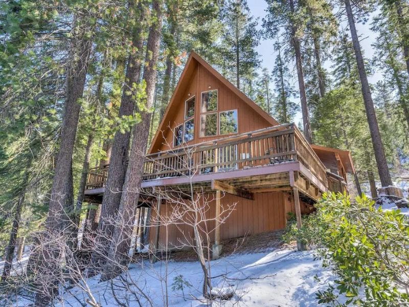 A charming A-frame cabin nestled among tall pine trees, with large windows and a spacious wooden deck.