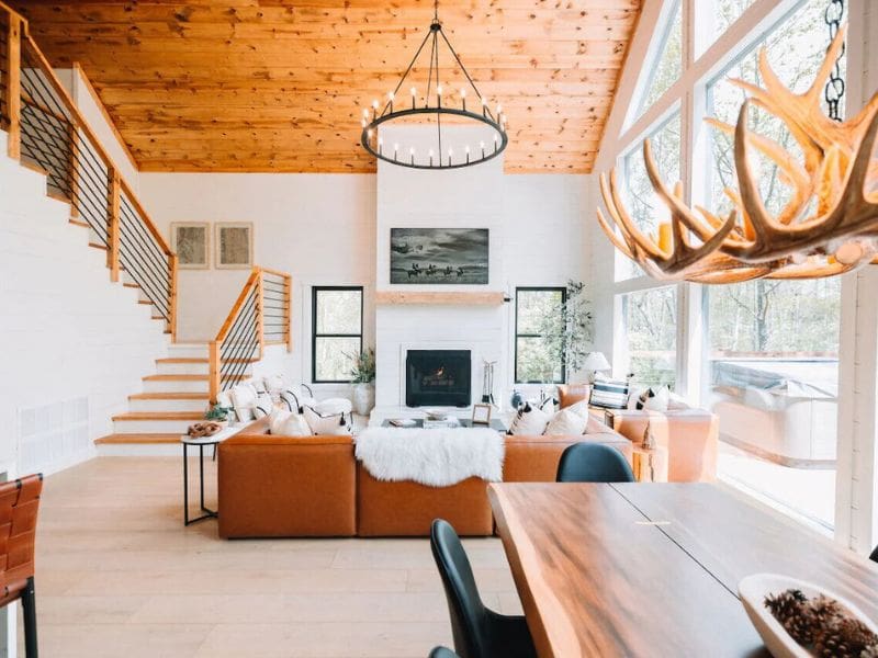 A modern, spacious cabin living room with large windows, a leather sectional sofa, a fireplace, and a grand antler chandelier. The room allows in ample natural light, highlighting wooden ceilings and an open staircase. Best cabin rentals in Hocking Hills, OH.