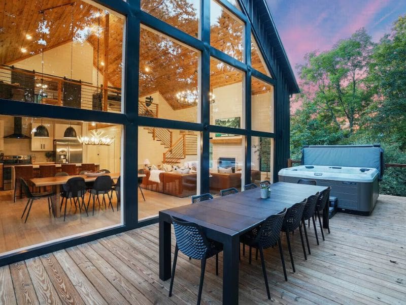 An outdoor dining area with a long table and chairs set on a wooden deck. Large glass windows reveal the cabin’s well-lit interior, and a hot tub is placed beside the deck. Best Hocking Hills cabin rentals.