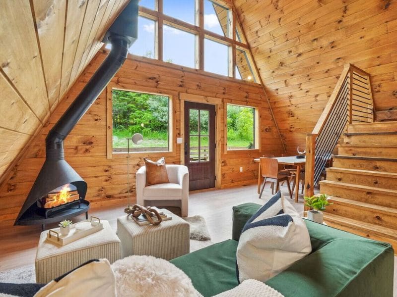 Interior of The Everwood A-Frame’s open living area with a Scandinavian-style fireplace, large windows overlooking the forest, and modern furniture. Cabins in Hocking Hills, OH.
