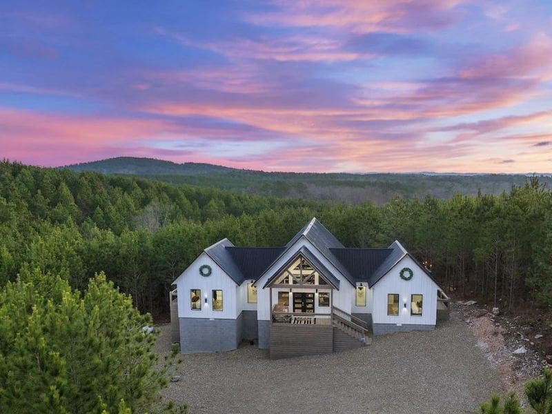 Exterior view of Heart & Hearth, a Broken Bow luxury cabin rental at dusk, showcasing its unique architectural design with expansive windows against a forested backdrop and a colorful sky.
