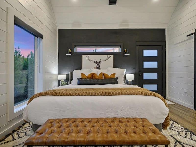 Master bedroom in Heart & Hearth cabin featuring a modern aesthetic with black walls, white linens, a leather bench, and a window.