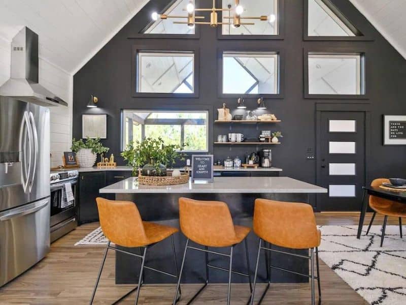 Modern kitchen inside Heart & Hearth, a Broken Bow luxury cabin, with dark walls, stainless steel appliances, an island with leather chairs, and upper windows.