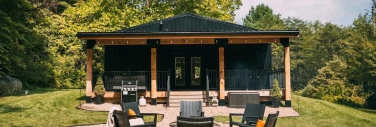 Front view of Ivy Cottage by Creative Cabins, featuring a modern black exterior with wooden accents and a spacious porch. The outdoor area includes comfortable seating around a fire pit and a grill, surrounded by greenery and trees. Luxury Cabin Rentals in Hocking Hills, Ohio.