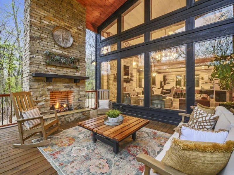 Outdoor patio with a brick fireplace at a Broken Bow 5-bedroom cabin rental with wooden rocking chairs and a view into the interior through large glass windows.