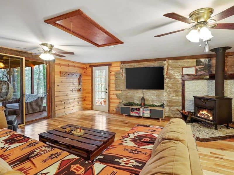 A cozy living room with a wood-paneled interior, ceiling fans, and a flat-screen TV mounted above a stone fireplace. The room is furnished with comfortable couches and features access to a screened-in porch. Dog-friendly cabin rental in Hocking Hills, OH.