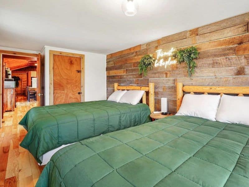 A bedroom featuring two wooden log-framed beds with green bedding, against a wood accent wall. Above the beds is a neon sign reading "There's No Place Like Home". Vrbo in Hocking Hills, OH.