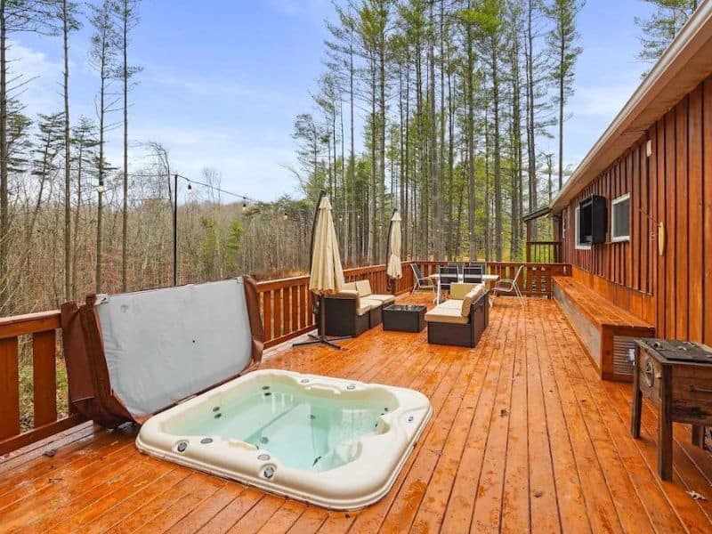 A spacious wooden deck surrounded by trees, with a large hot tub, outdoor seating area, and umbrellas for shade. Where In The Woods cabin rental, Hocking Hills, OH.