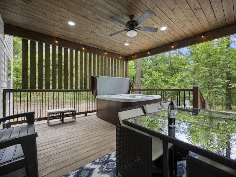 Covered deck area of a Broken Bow cabin rental with a hot tub, outdoor seating, and a forest backdrop.