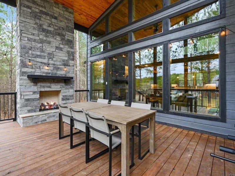 Outdoor dining area with a stone fireplace on the deck of a Broken Bow Vrbo cabin rental, including a wooden table with seating and forest views through floor-to-ceiling windows.
