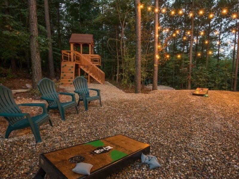 Outdoor recreational space at a large Broken Bow cabin rental with Adirondack chairs, string lights, and a children's playset in the background.