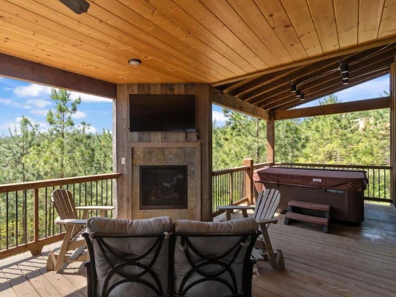 Deck area of a Broken Bow cabin rental featuring a stone fireplace, outdoor TV, and a hot tub, perfect for entertaining and relaxing in nature.