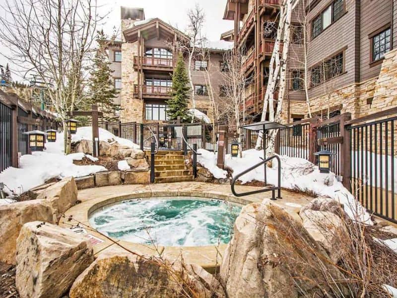 Outdoor hot tub at Arrowleaf Penthouse Vrbo rental in Park City, surrounded by rocks and snow, with the multi-story building in the background.