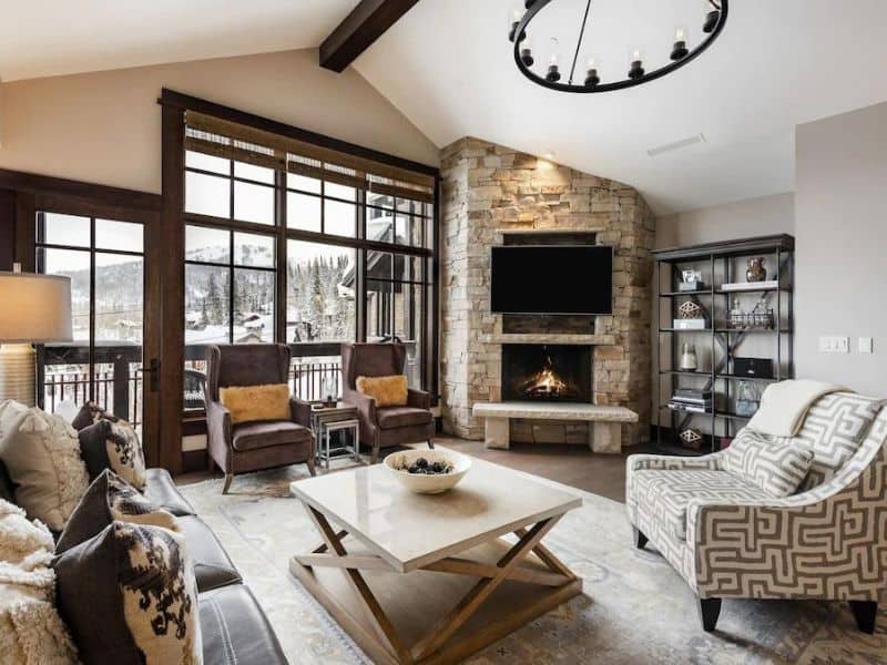 Stylish living room in Park City's Arrowleaf Penthouse, with large windows with snowy mountain views, a stone fireplace, a wall-mounted TV, and comfortable seating.