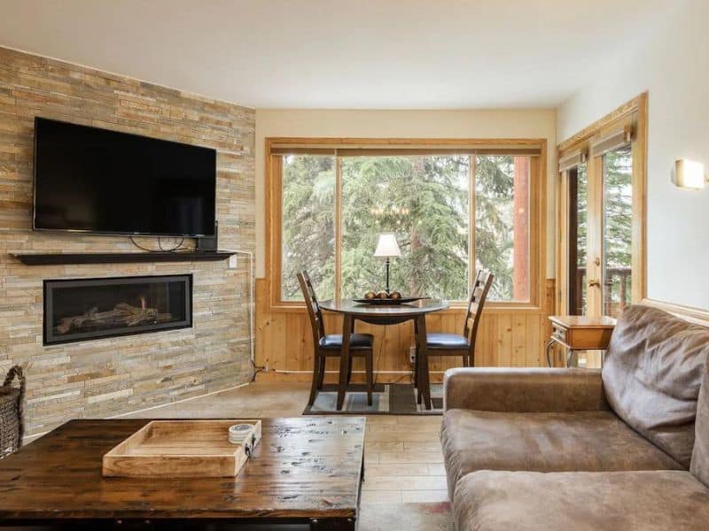A cozy living room in a Park City ski in ski out rental with a stone fireplace, modern flat-screen TV, and a dining area overlooking pine trees through large windows.
