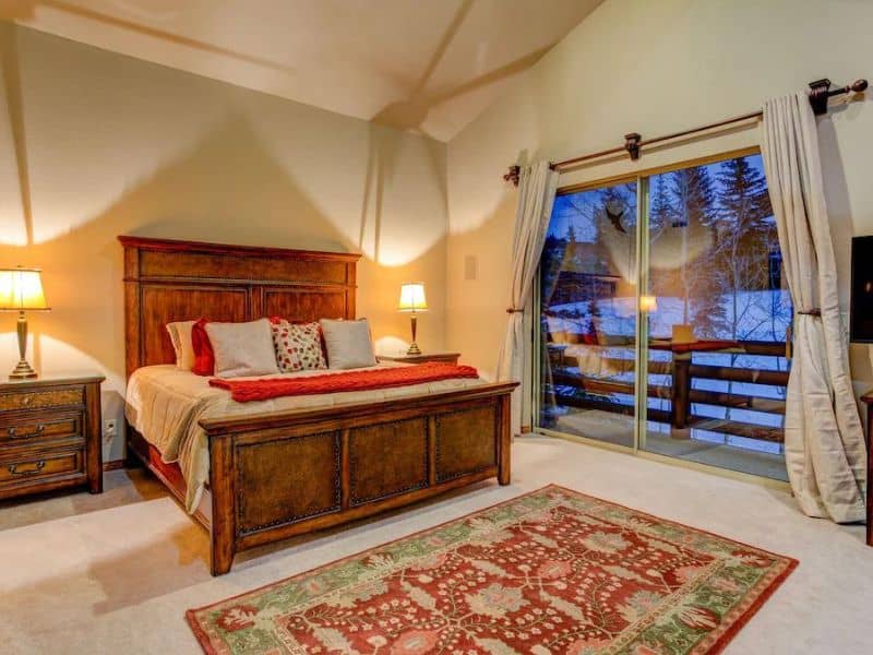 Master bedroom in Sterling Vista Vrbo in Park City with a large wooden bed with elegant bedding, two bedside lamps, and sliding glass doors that lead to a private balcony with snowy mountain views.