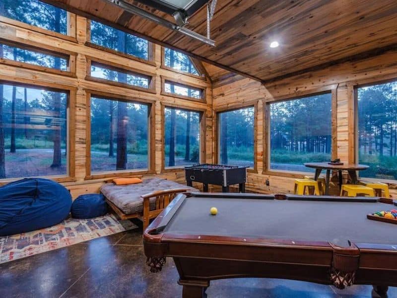 Game room with a pool table and large windows overlooking the forest at 'Skip-A-Rock,' a family-friendly 5-bedroom Broken Bow cabin rental.