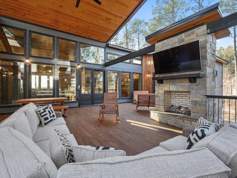 Outdoor lounge area of a Broken Bow luxury cabin rental with plush seating, a stone fireplace, and expansive glass windows overlooking the woods.