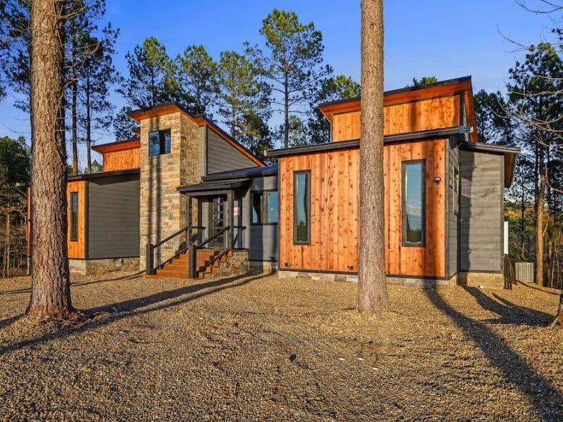 Exterior view of a modern 5-bedroom cabin in Broken Bow with a unique mix of wood and stone finishes, surrounded by tall pine trees.