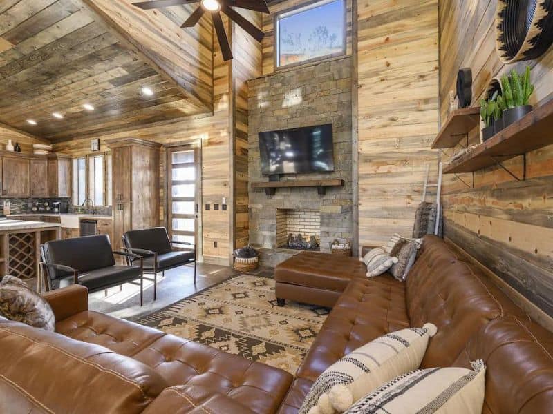 Interior of a cozy 5-bedroom Broken Bow cabin rental featuring a spacious living area with leather couches, a stone fireplace, and rustic wooden walls.