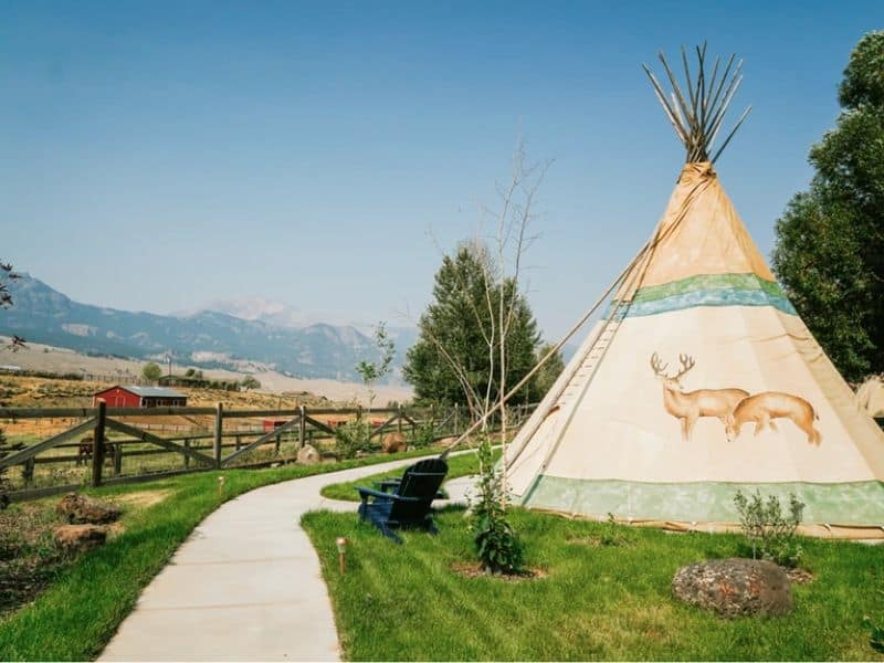 Traditional tipi with decorative elk artwork set against a backdrop of Montana's mountains, part of a glamping experience at Yellowstone Tipis.