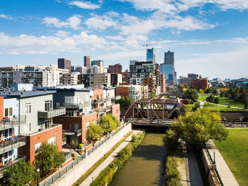 Aerial view of downtown Denver with modern apartment buildings along a canal, a historic iron bridge, and a backdrop of skyscrapers under a bright blue sky with scattered clouds. Part of a "Denver to Grand Canyon Road Trip Itinerary" blog post.