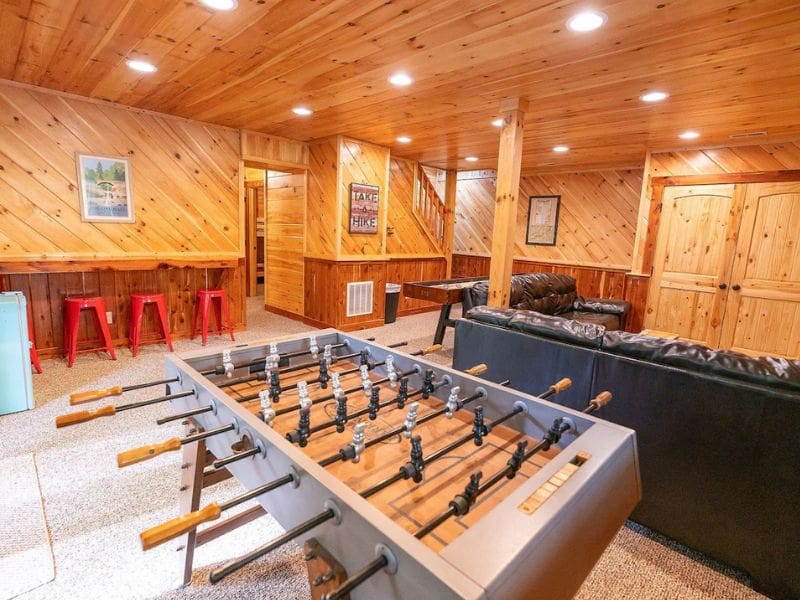 A basement game room with wood-paneled walls, a foosball table, and a cozy seating area. Red stools line the wooden bar. Redwood Lodge - a pet-friendly cabin rental in Hocking Hills, OH.