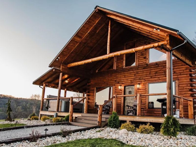 A log cabin with large windows and a spacious wooden porch. The cabin is surrounded by well-maintained landscaping and rock beds, set against a backdrop of rolling hills. Shurtz Overlook in Hocking Hills, OH.