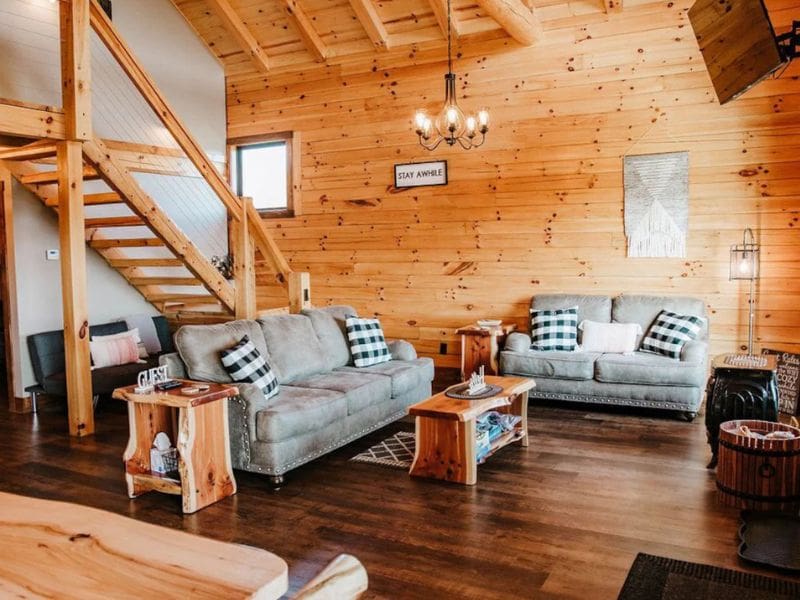 Living room inside "Shurtz Overlook," a log cabin rental in Hocking Hills, OH. The room features warm wooden walls and high vaulted ceilings, with two comfortable gray sofas adorned with plaid pillows. A wooden staircase leads to an upper loft. Dog-friendly retreat in Hocking Hills, Ohio.
