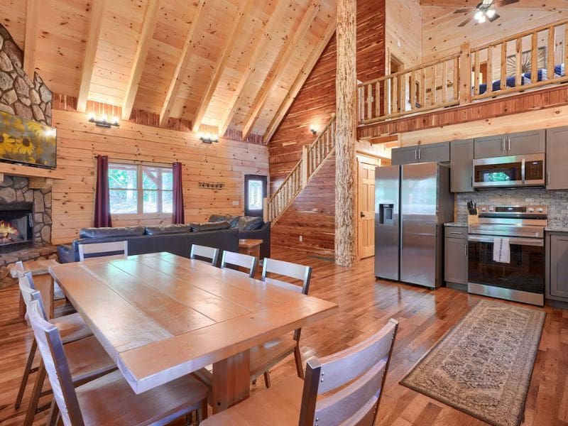 A rustic log cabin interior with vaulted wooden ceilings and exposed beams. The open-plan space features a dining area with a large wooden table and chairs, a modern kitchen with stainless steel appliances, and a cozy living area with a stone fireplace and TV mounted above it. An upper loft is visible. Star Ridge Cabin, a pet-friendly cabin rental in Hocking Hills, OH.