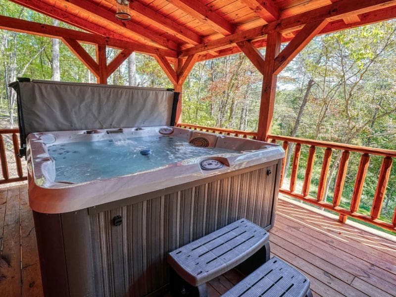 A covered outdoor deck featuring a hot tub with wooded surroundings in the background. Star Ridge Cabin in Hocking Hills, OH.