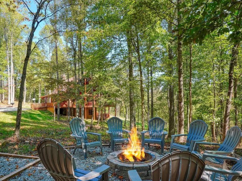 An outdoor seating area with several Adirondack chairs arranged in a circle around a fire pit. The fire is surrounded by tall trees, with a cabin visible in the background.