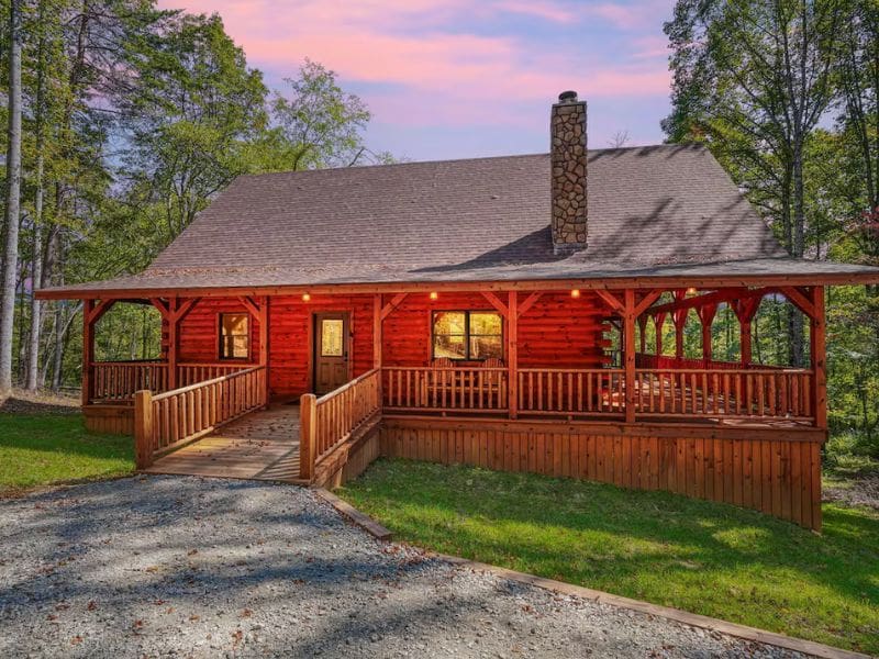 Front view of a pet-friendly log cabin rental in Hocking Hills, OH with a spacious, wraparound porch. The cabin's exterior features stone accents on the chimney, surrounded by a wooded area with evening lighting.