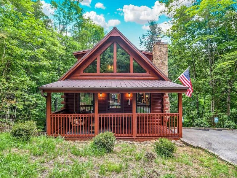 Exterior of Pigeon Forge rental cabin, with classic log cabin construction, American flag, and wrap around front porch, and large windows. surrounded by green forest.