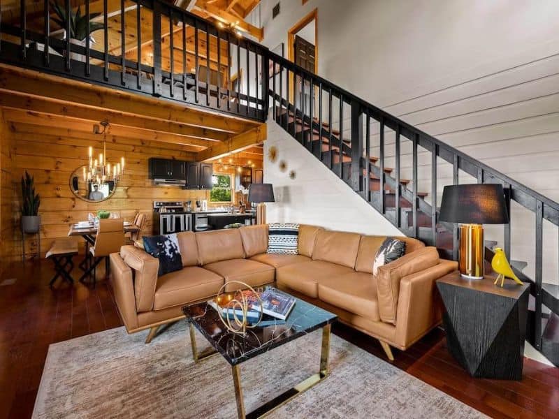 Living area of Pigeon Forge vacation cabin, with mid century modern styling, brown leather L- shaped couch, and dining room behind with modern chandelier.