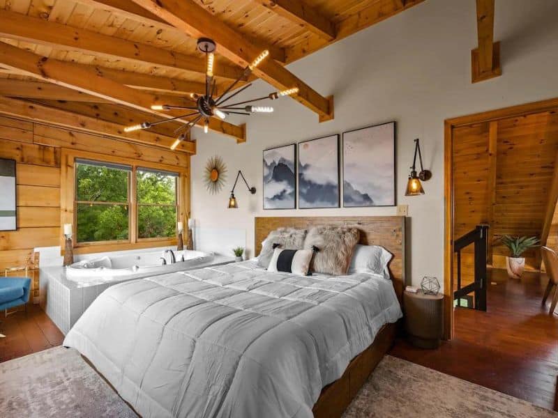 Master bedroom on Pigeon Forge vacation cabin, with exposed wooden beams on the ceiling, a jacuzzi tub in the corner, and mid century modern rustic decor. Views of forest.