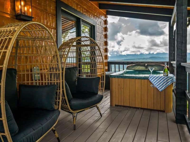 Two birdcage chairs and a jacuzzi tub perch on the balcony of Pigeon Forge vacation cabin, with a surrounding mountain view.