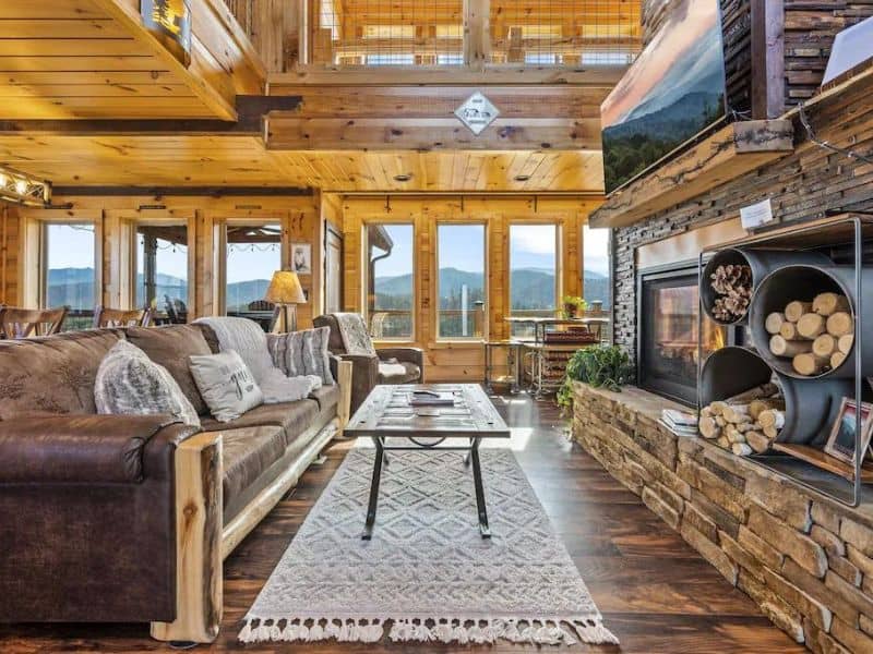 Rustic modern room in cabin with couch, coffee table with rug, and modern stone fireplace with feature built in to hold wood