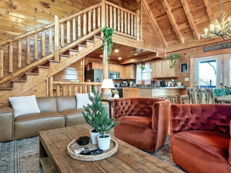 Living room of Pigeon Forge rental cabin, featuring plush red midcentury style chairs and evergreen accents, wood paneling throughout, kitchen and dining area beyond, with an antler chandelier.