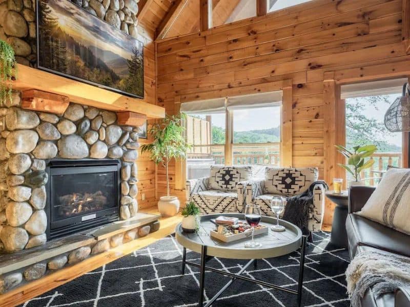 Living room with modern black and white decor, wood paneling floor to ceiling, and a large river rock wood burning fireplace, with views of the forest.