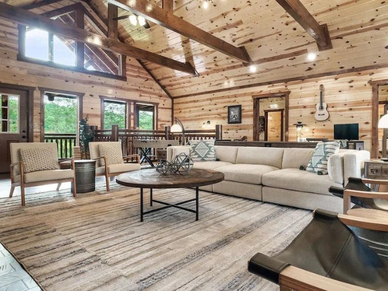 Living area of Pigeon Forge vacation cabin, with light neutral tones and scandinavian style details. Angeled wood ceiling has exposed beams, and large windows look out to the forest.