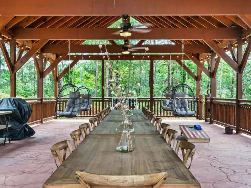 Rooftop dining area of Pigeon Forge rental home. An exposed beam covered porch houses a Large wooden dining table and chairs, with 4 hanging birdcage chairs and accent lighting.