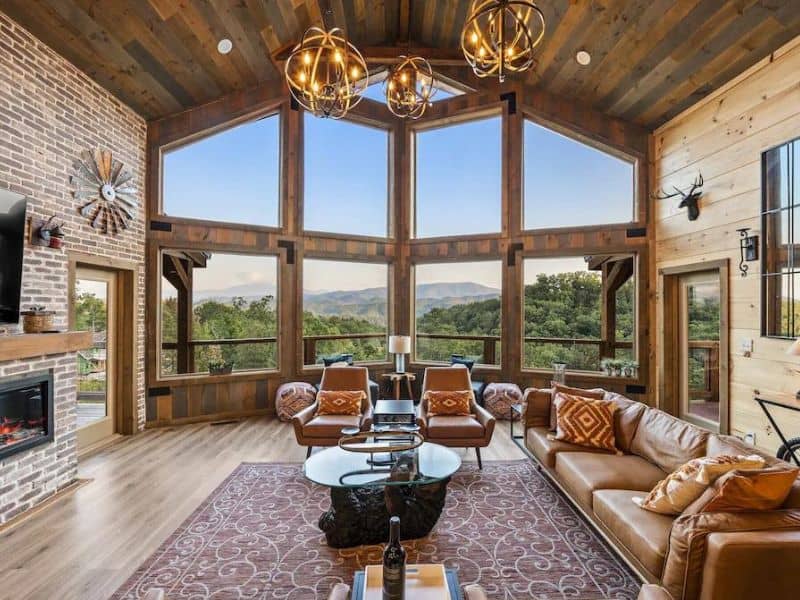 Living area of Pigeon Forge cabin, with vaulted ceilings, fire place, and mid-centry style detailing. Large windows allow views of forested terrain.