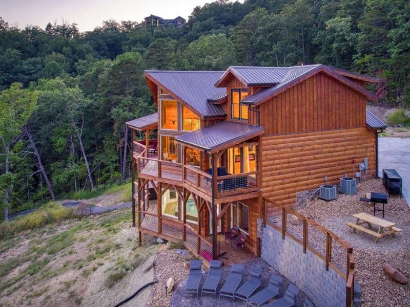 Exterior shot of Pigeon Forge Cabin rental, with dual level wrap around porches and outdoor seating areas. Log cabin style paneling and views of forested mountains.